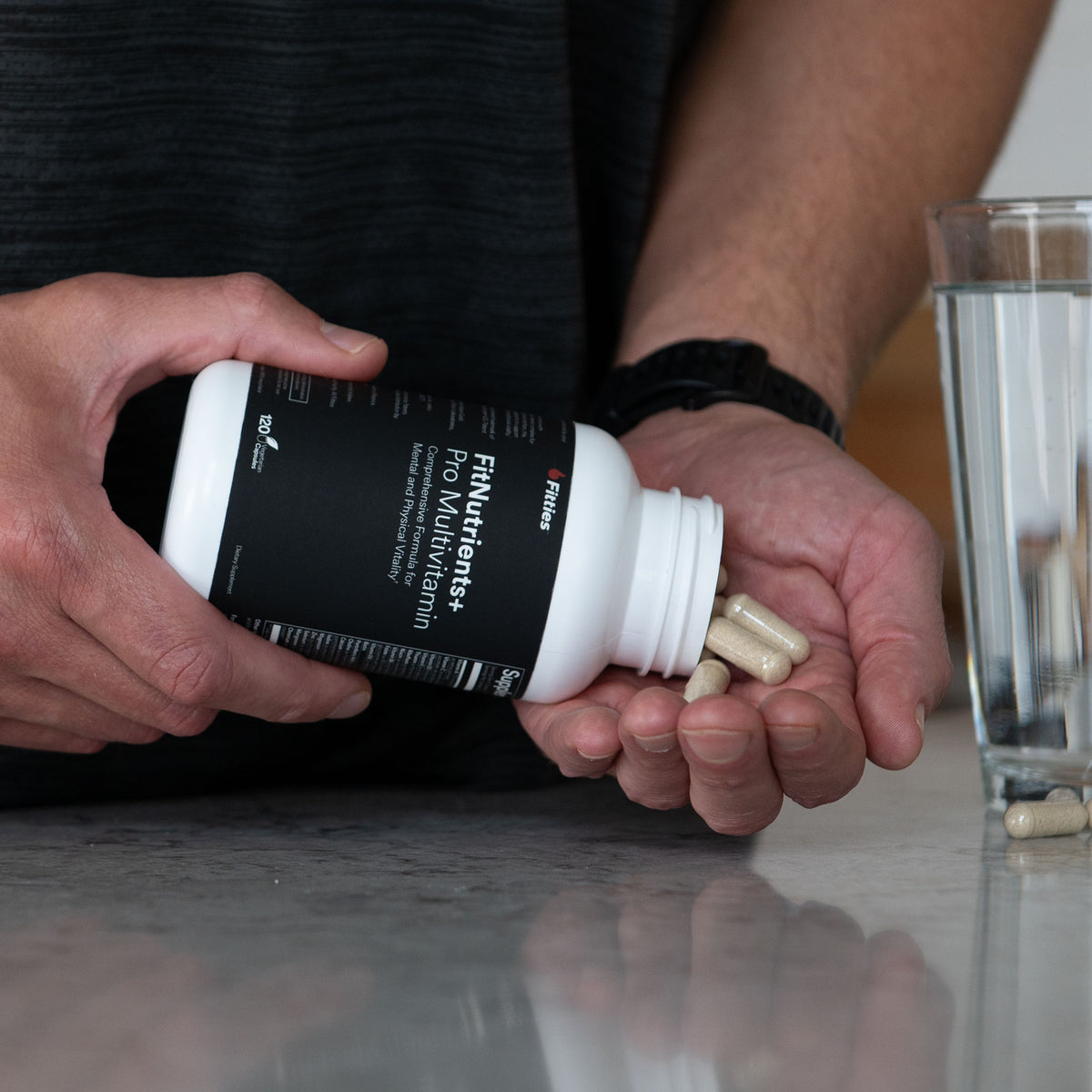 FitNutrients+ capsules poured from the bottle into a hand beside a glass of water