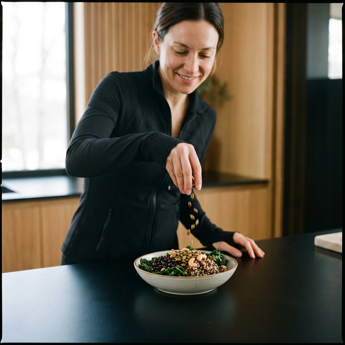 Woman sprinkling pumpkin seeds over a grain bowl with greens, black beans and cashews