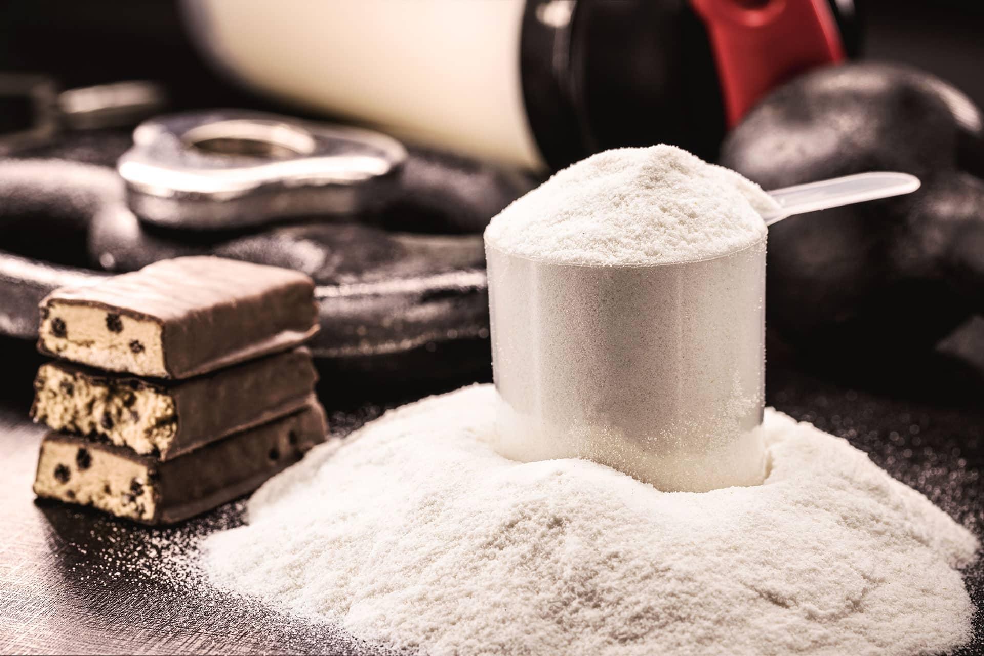 Closeup of scoop of vanilla protein powder with meal replacement bars, shaker bottle, and weight training equipment in background