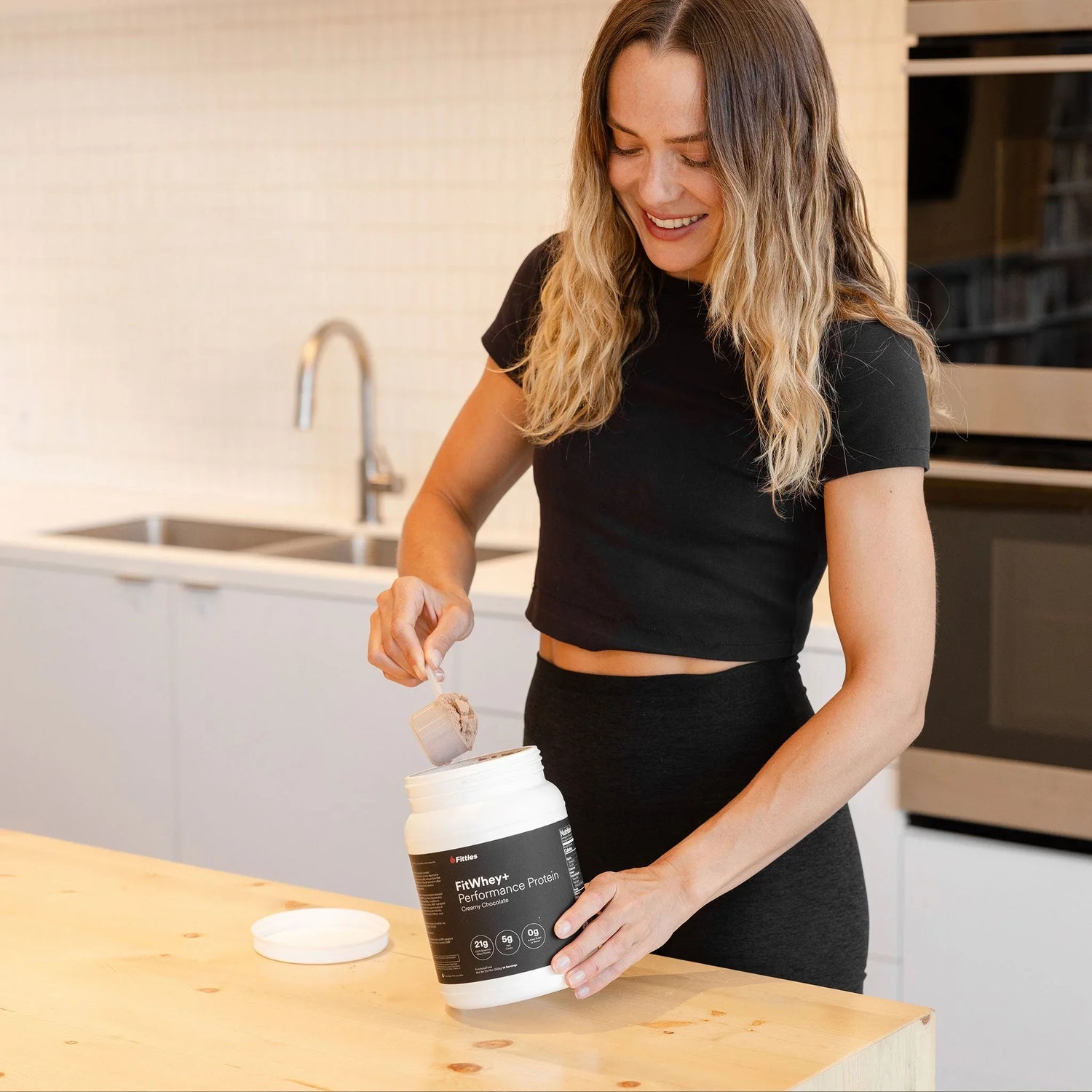 Woman in a kitchen scooping FitWhey+ grass-fed whey protein powder from the tub for a muscle gain shake
