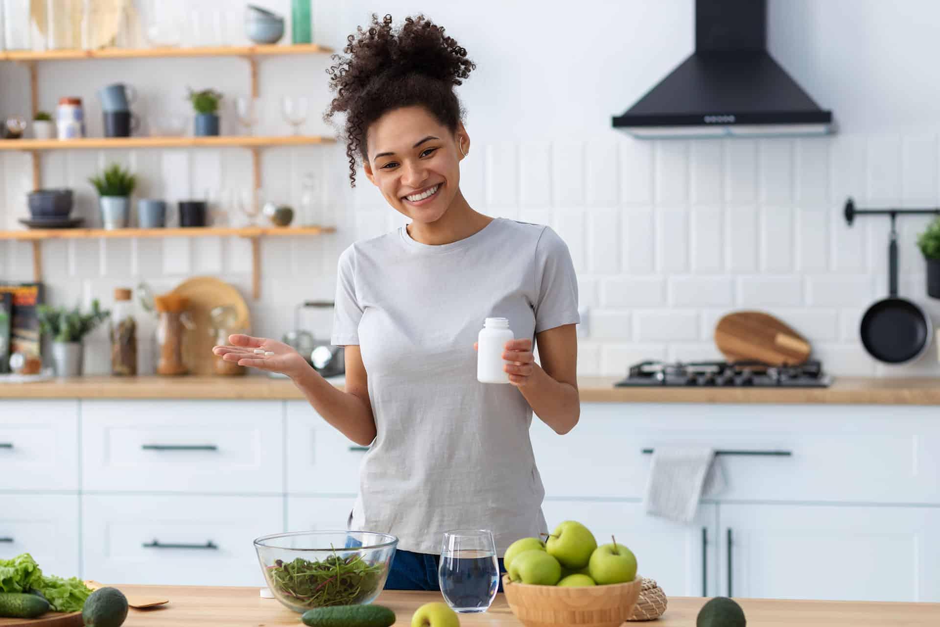 Female smiling while holding supplement capsules and bottle in hands in kitchen