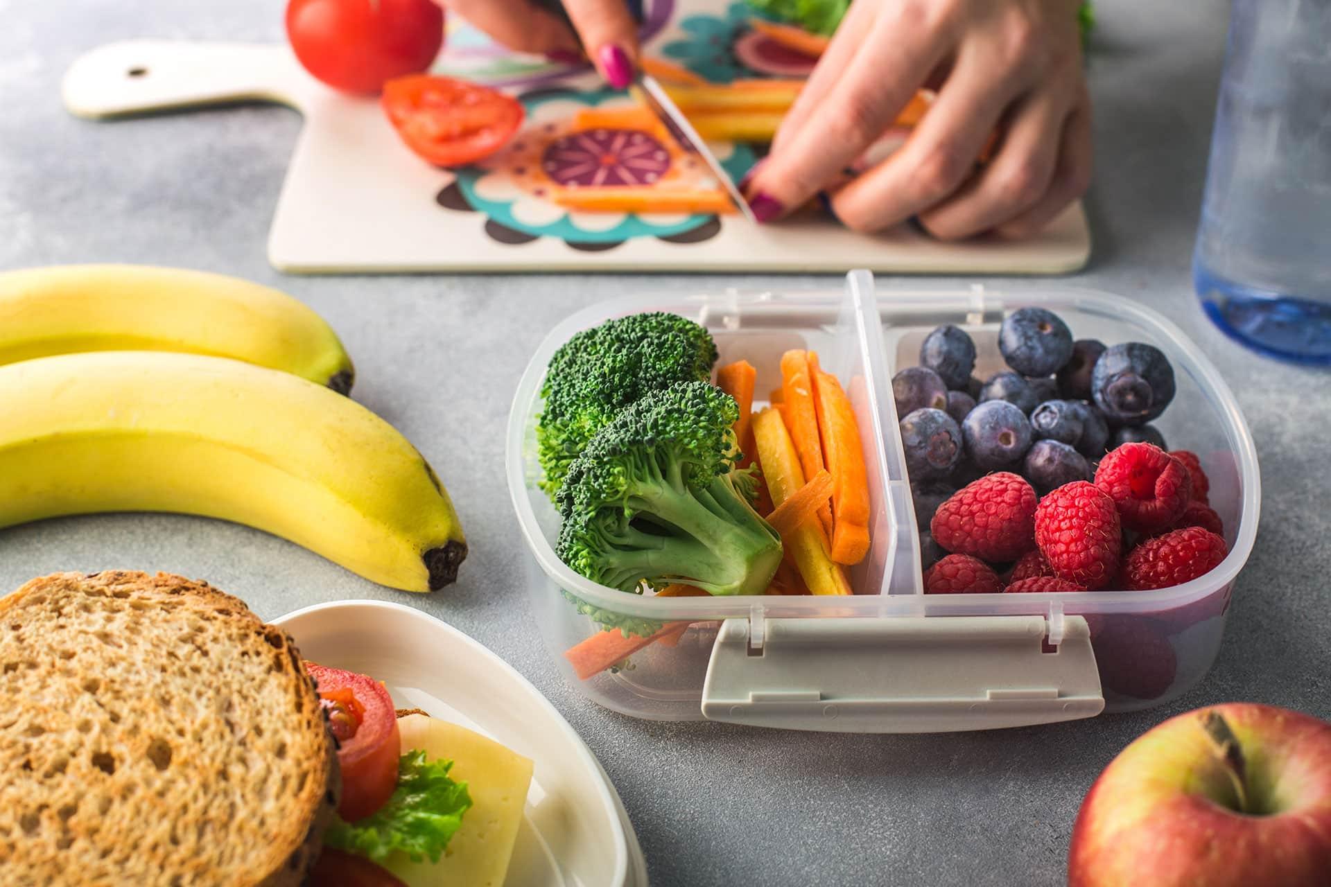 Closeup of snack container filled with berries and vegetables with female cutting veggies in the background