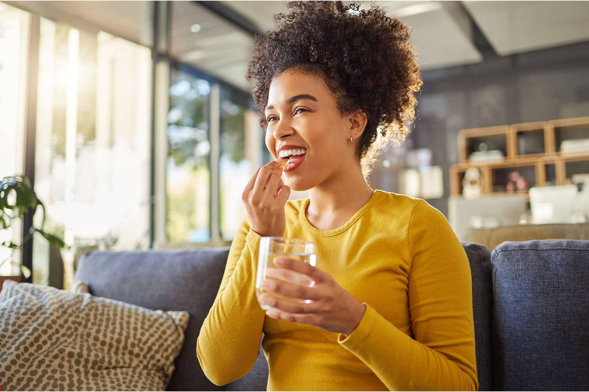 Female taking gel capsule supplement with water on sofa